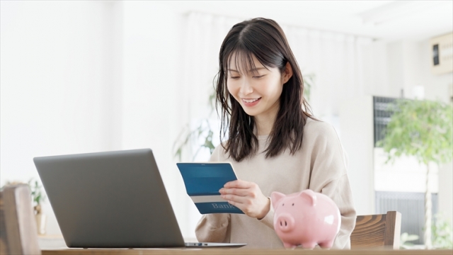 通帳を手にして微笑む若い母親とパソコンと貯金箱