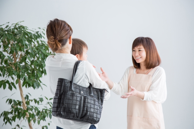 母子家庭のお母さんが子どもを抱っこして保育士に登園をお願いしている様子。保育園での温かいやりとりが伝わるシーン。