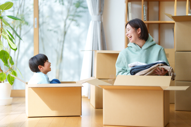 引っ越しの段ボールに囲まれながら笑顔で話す母親と子ども。母子家庭の新生活を明るく始める様子を表すイメージ。