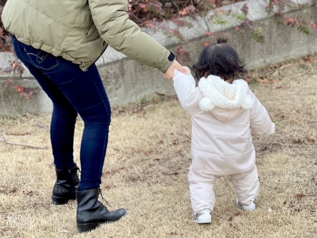 母子家庭の母親が幼い子どもの手を取り、屋外で歩く様子。支援団体や地域の助けを受けながら、前向きに子育てをしている場面。